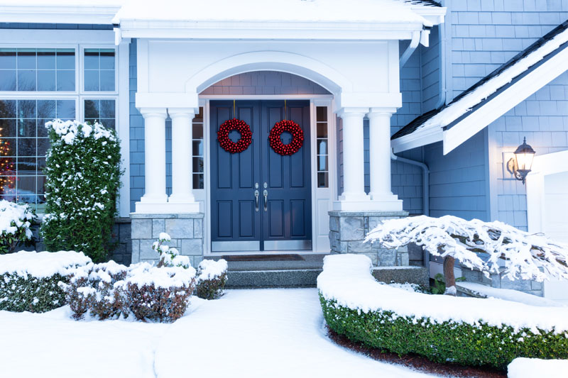 Close up of blue double doors of house with red wreaths on it with house covered in snow